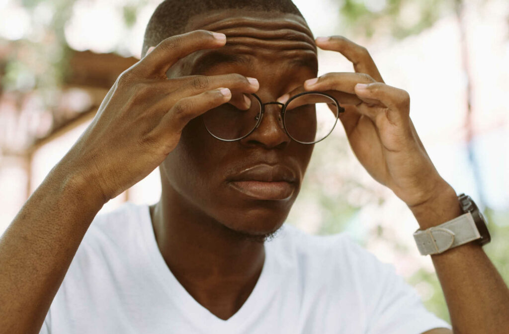 A young man sitting outside with his eyes closed and rubbing his eyes with both hands behind his glasses.
