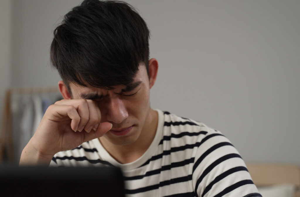 A young man squinting and rubbing his right eye while using a computer.