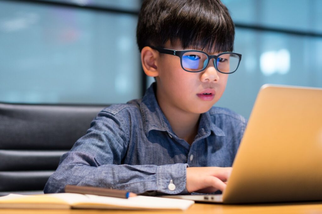 A young boy wearing a pair of blue light glasses while working on a laptop.