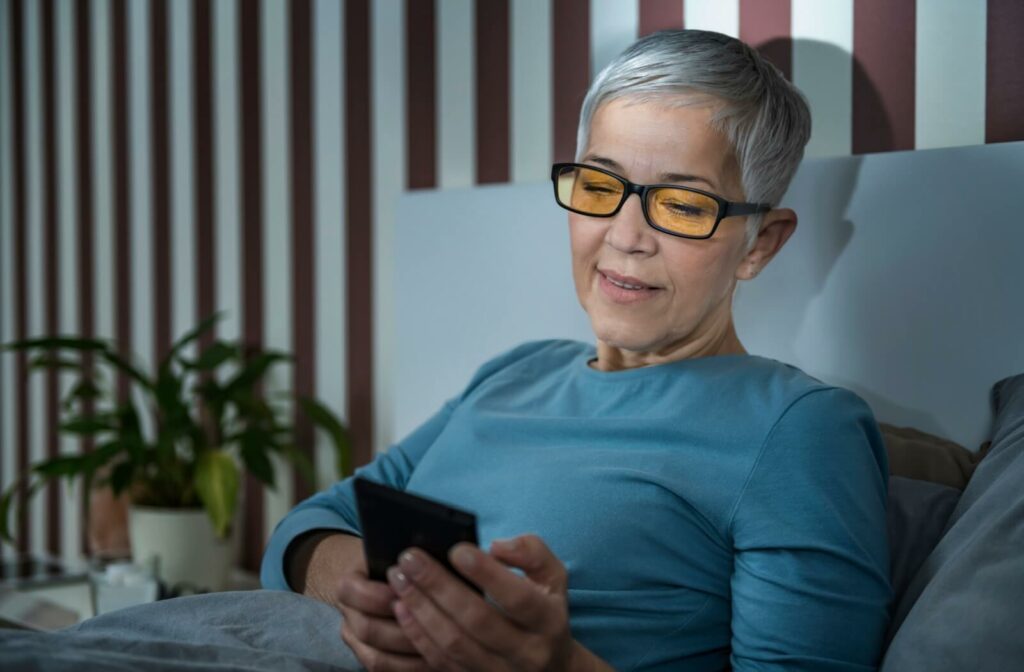 An older adult relaxes in bed at night reading on an electronic device with the aid of blue-light eyeglasses