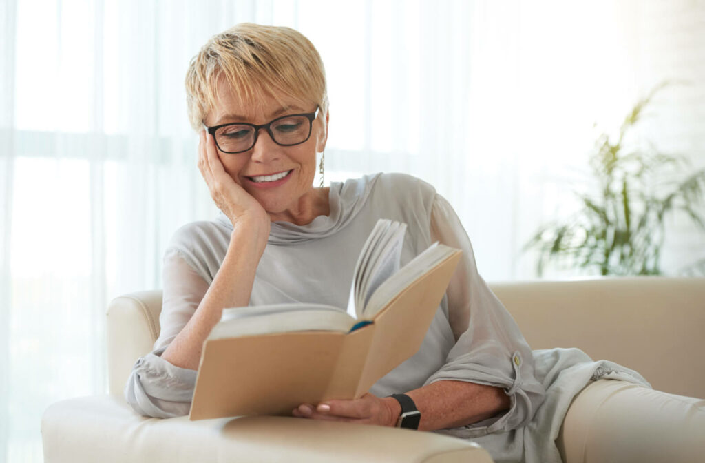 An older adult leaning on the arm of their couch reading a book and smiling due to their new reading glasses.