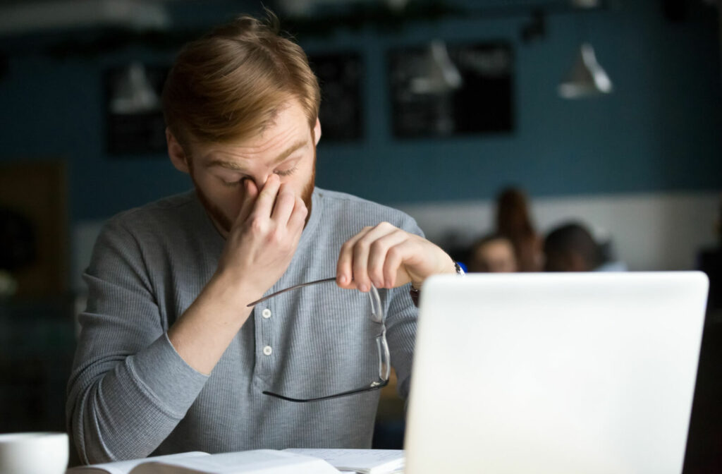 A young adult working at their laptop, taking off their glasses and rubbing their eyes due to discomfort.