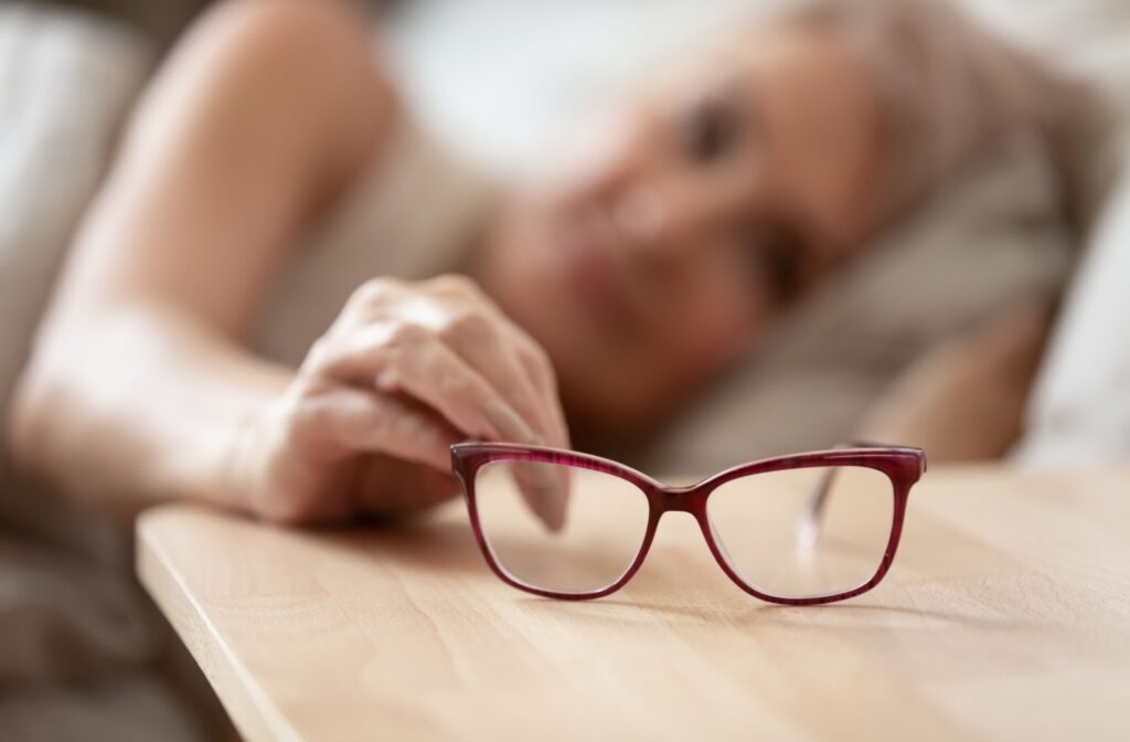 A person lying down in bed, reaching for their red glasses as they wake up.