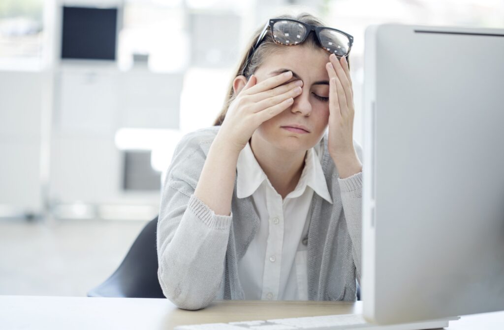 Young woman rubbing her eyes in front of a computer screen, showing signs of digital eye strain or fatigue.