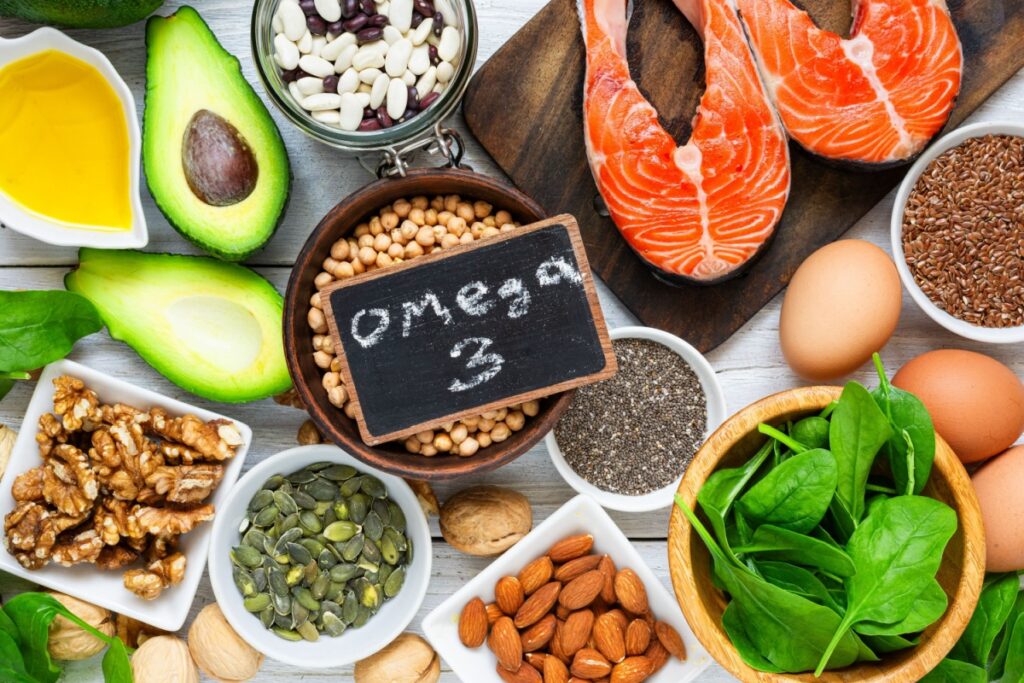 Assortment of omega-3 rich foods including salmon, avocado, walnuts, almonds, chia seeds, flaxseeds, eggs, and leafy greens arranged on a tabletop.