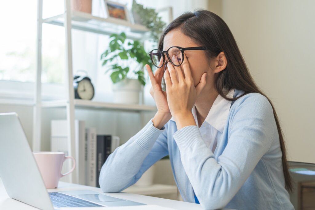 Woman wearing glasses rubbing her eyes while sitting at a laptop desk, suggesting eye strain or digital eye fatigue during computer work.
